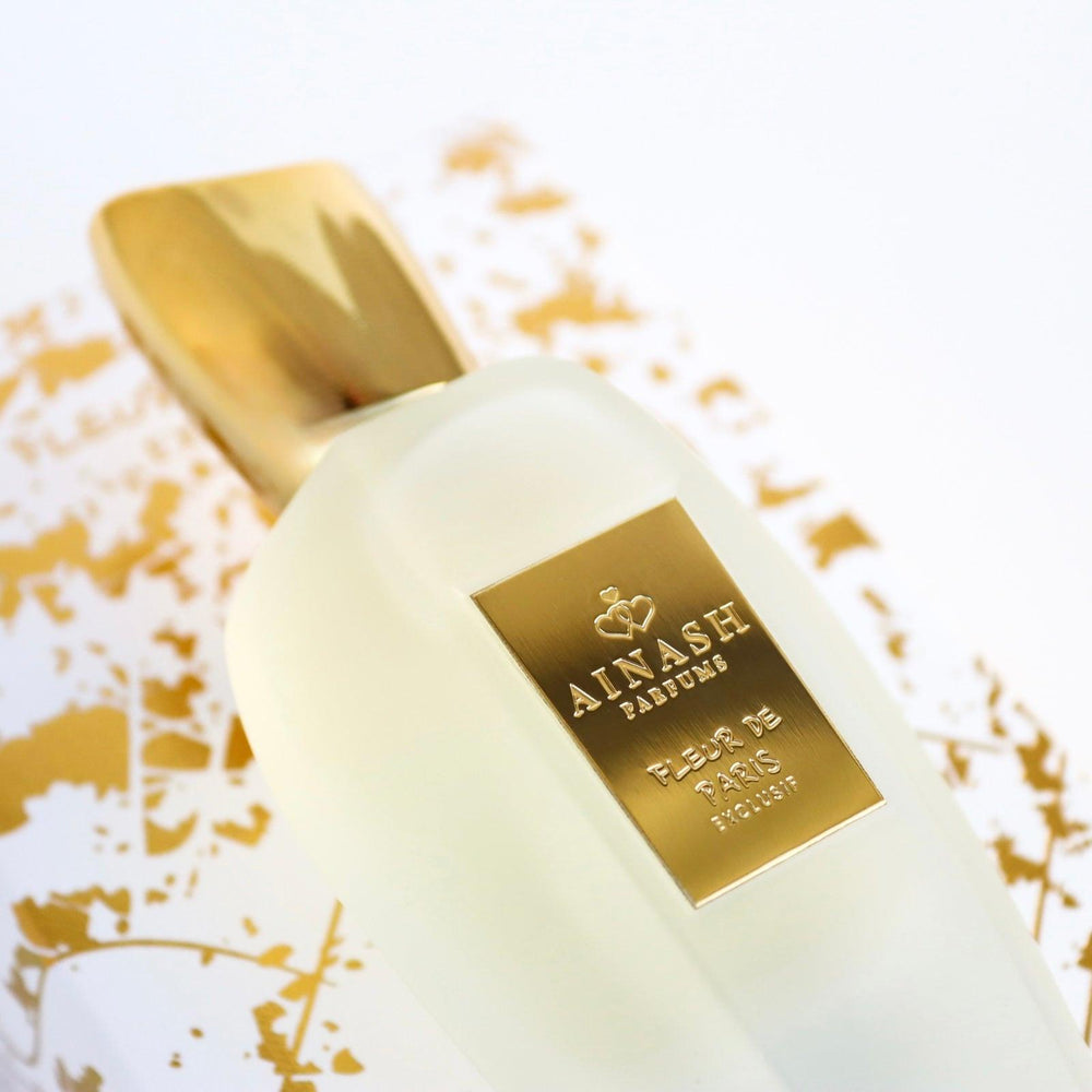 "Fleur de Paris Exlusif extrait de parfum by Ainash Parfums — frosted white bottle with gold label and cap resting on gold-detailed packaging"